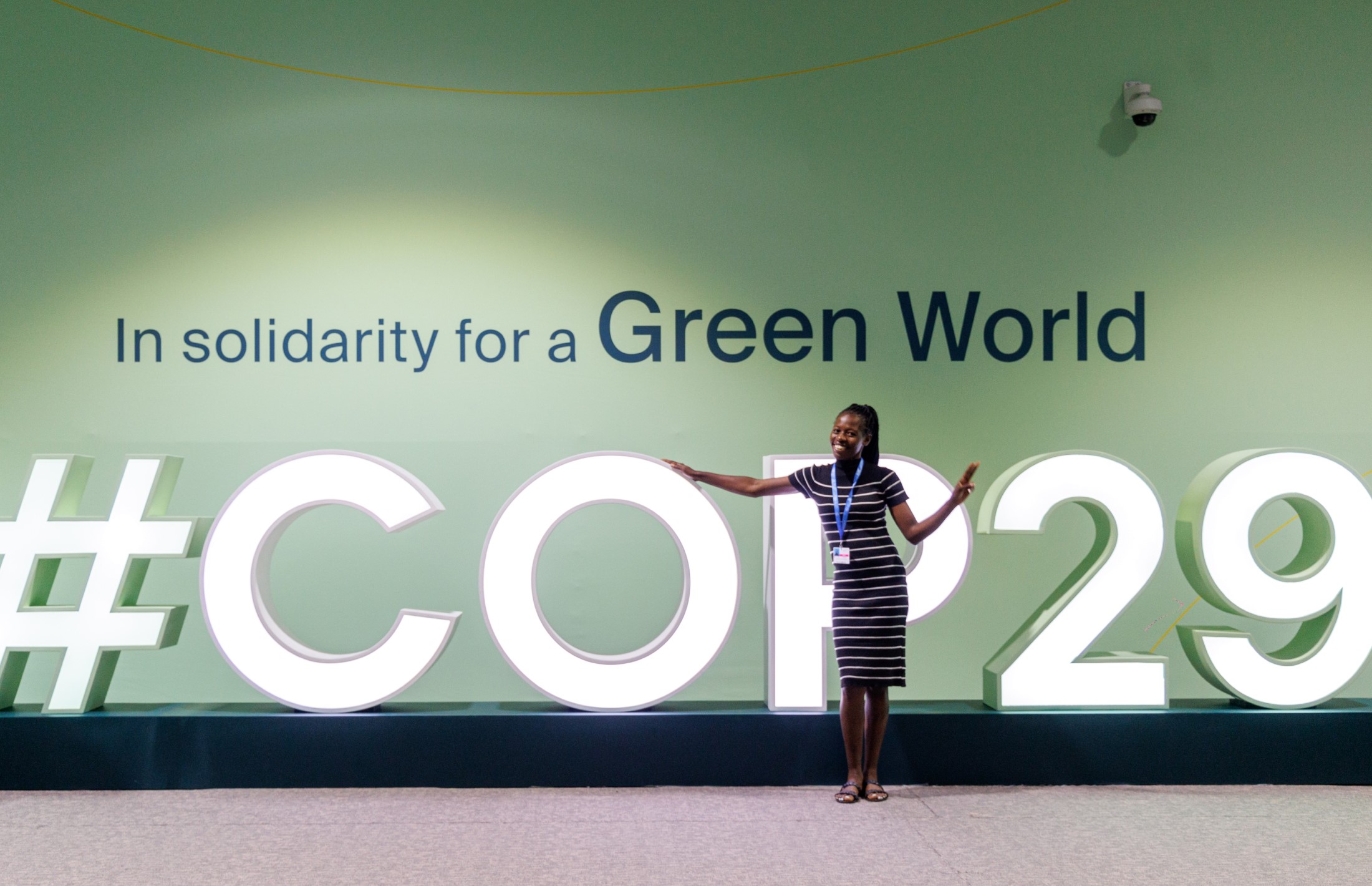 A woman standing in front of an installation that says #COP29. On the wall, behind the installation, it's written: "In solidarity for a Green World.".