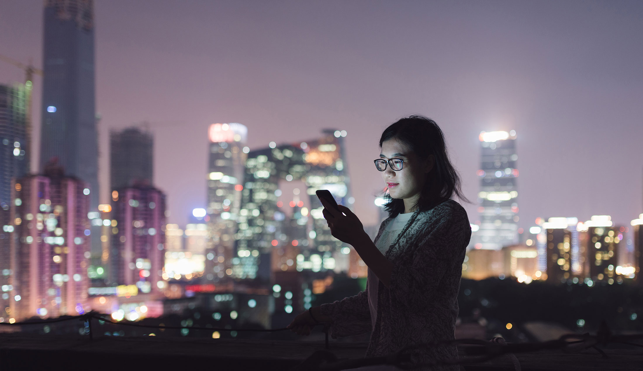 Kvinna står framför skyline med höghus nattetid och läser på sin mobil.