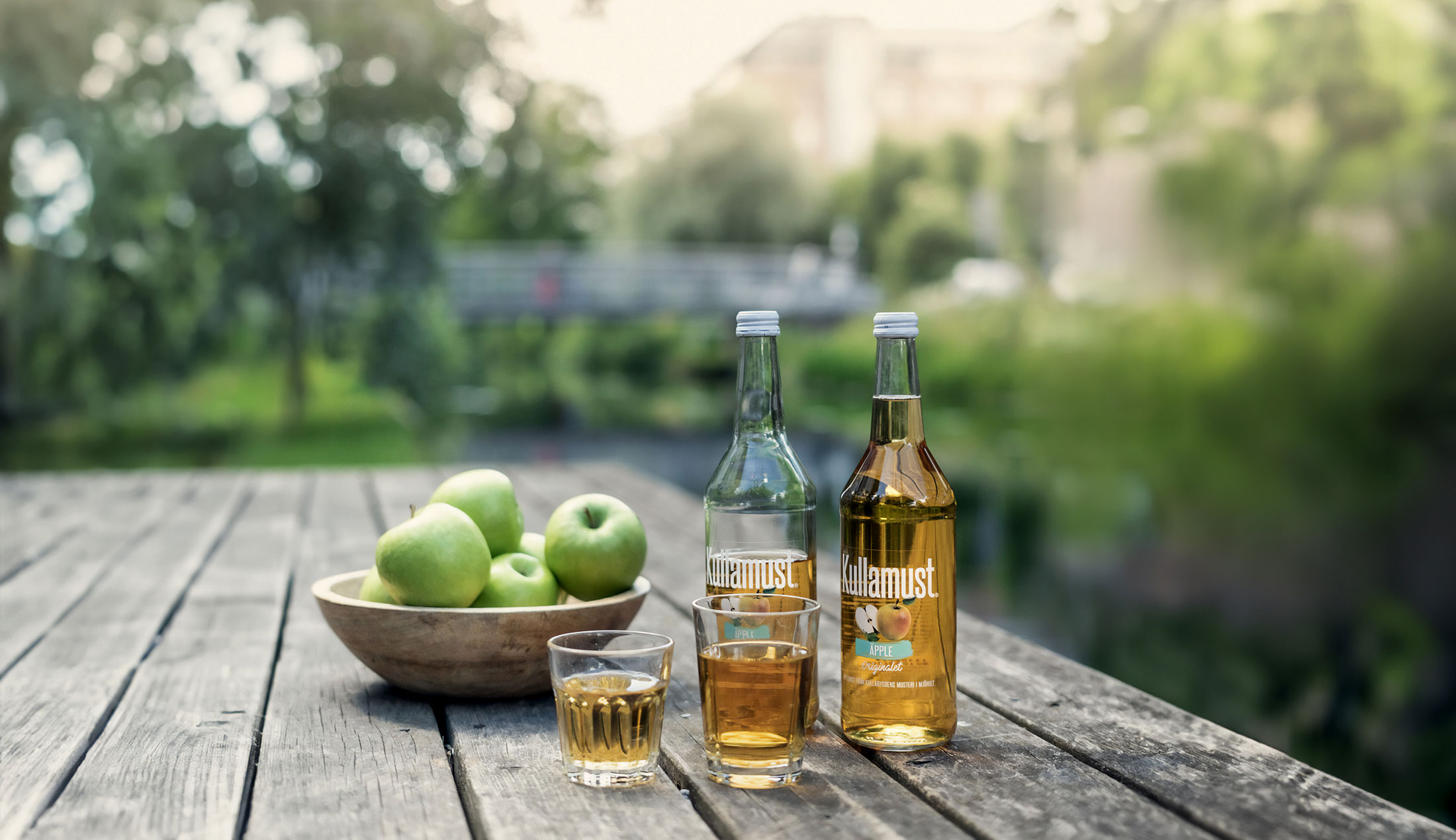 Två glas med äppelmust står på ett bord framför en skål med äpplen och två flaskor kullamust.