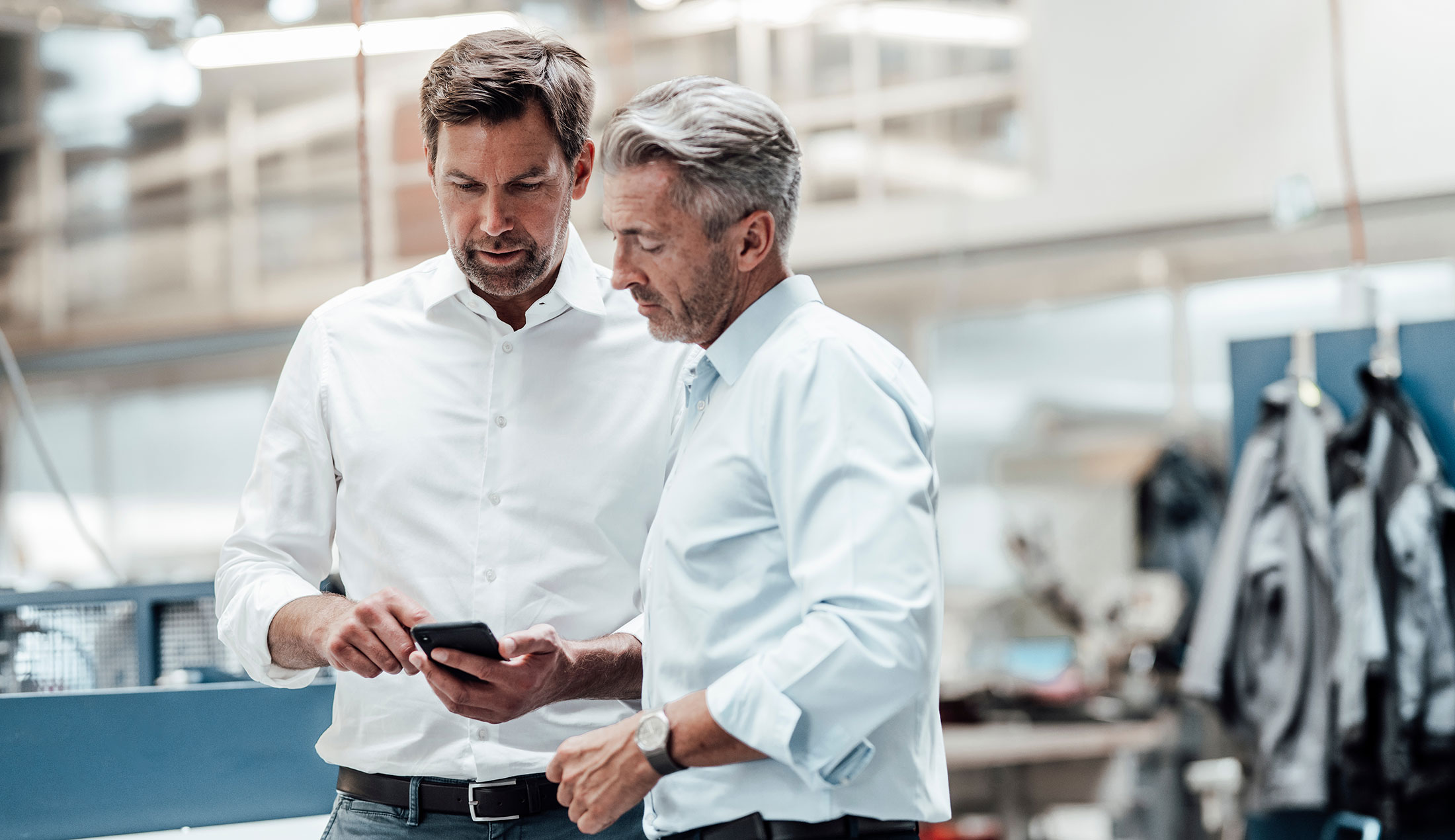 ChatGPT sade: Two men standing in a bright industrial setting, looking at one of the men’s mobile phone.