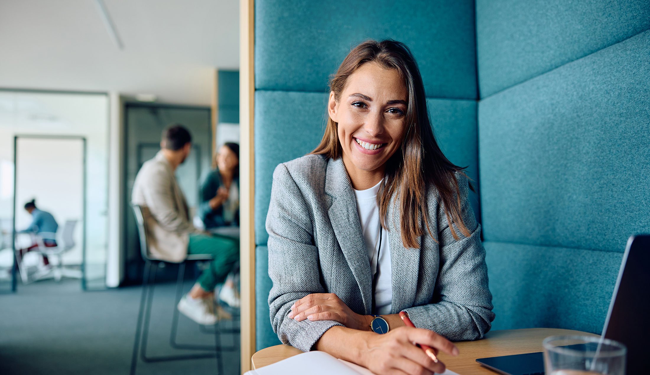 Leende kvinna vid ett skrivbord på ett kontor med andra personer i bakgrunden.