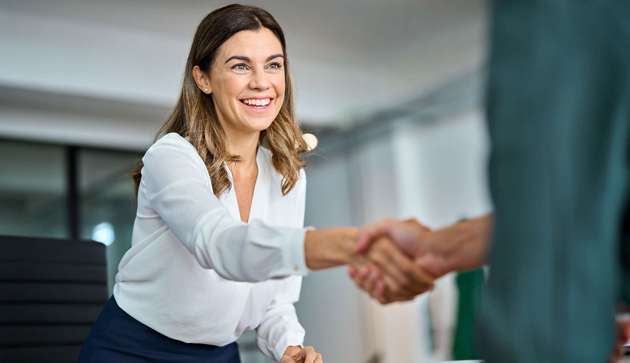 A businesswoman is half-standing by her desk, shaking hands with a man seen from behind.
