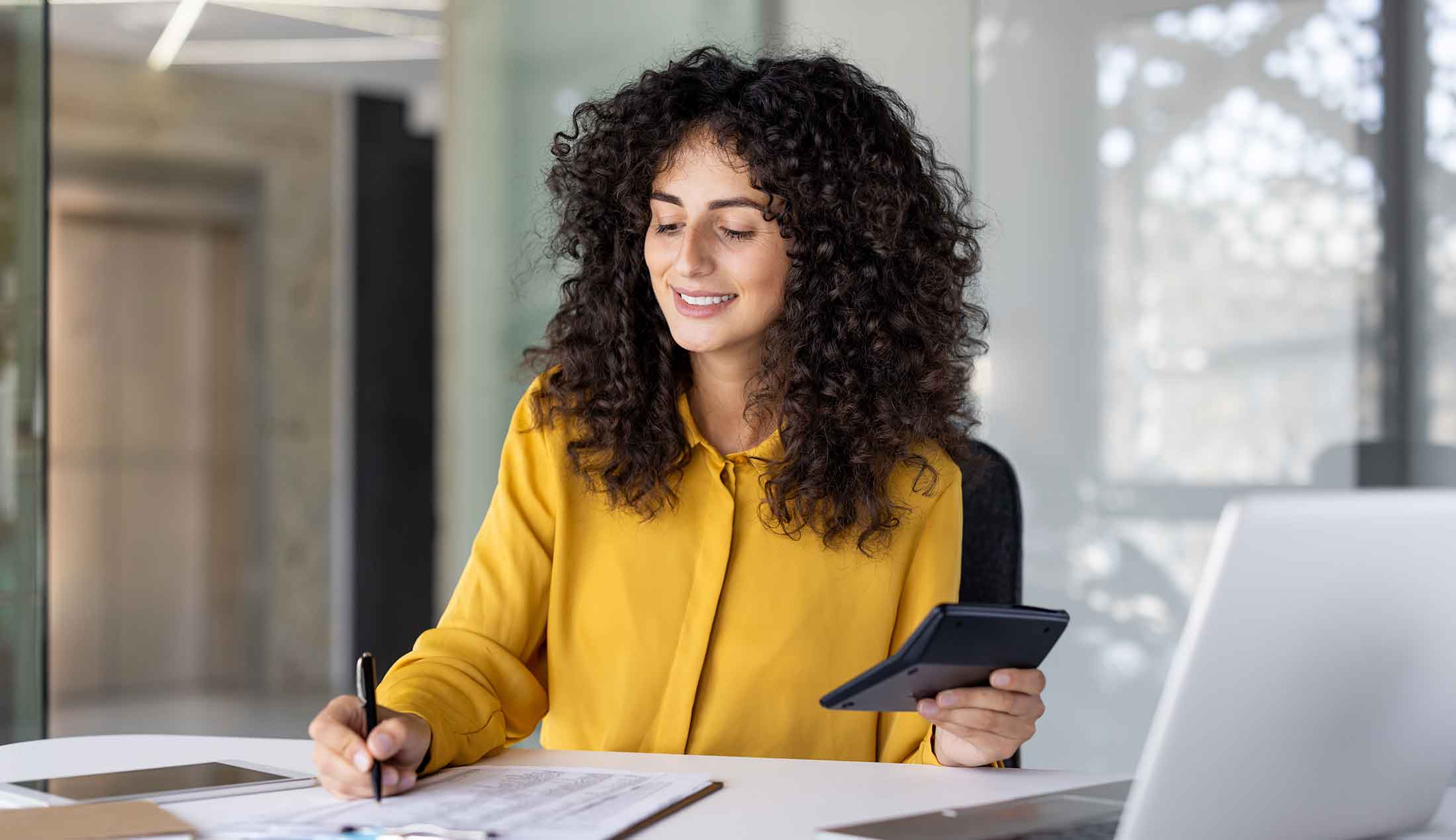 A woman is sitting at a desk, writing something on a piece of paper while holding a calculator in her other hand.
