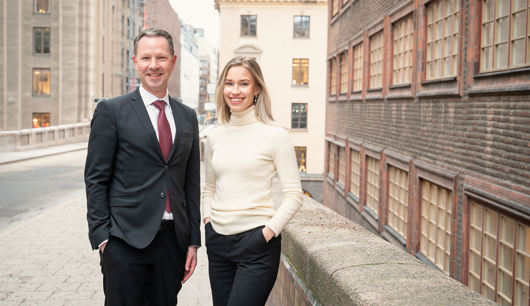 A man and a woman are standing on a city street, looking towards the camera.